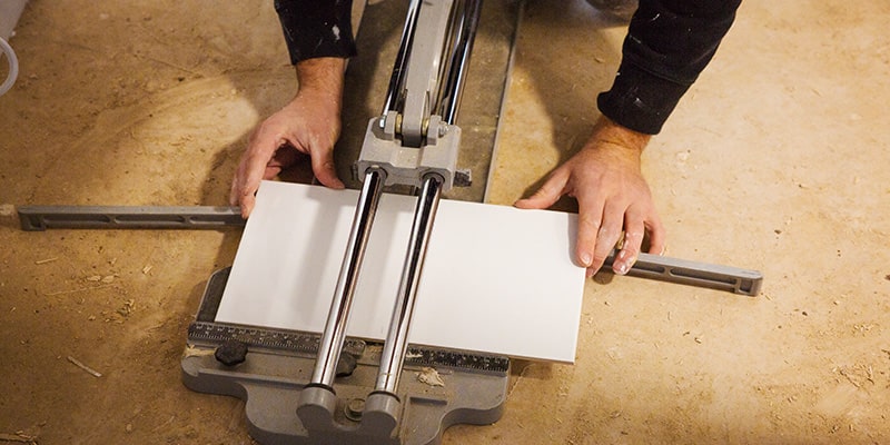 Builder kneeling down to cut a white procelian tile with a manual tile cutter.