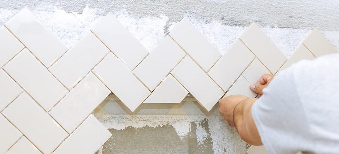 Worker tiling wall with while subway tiles in a herringbone pattern.